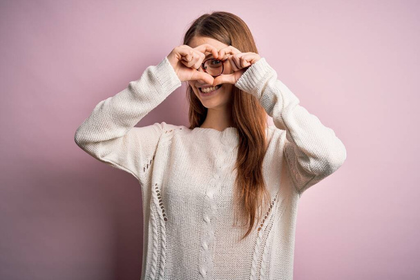 Young beautiful redhead woman wearing casual sweater and glasses over pink background Doing heart shape with hand and fingers smiling looking through sign - Zdjęcie, obraz