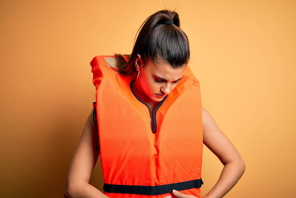 Fiatal gyönyörű barna nő visel narancssárga biztonságos mentőmellényt sárga háttér kézzel a gyomron, mert emésztési zavar, fájdalmas betegség rossz közérzet. Ache-koncepció. - Fotó, kép