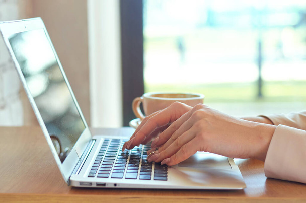 Młode kobiety z jasnoróżowym manicure wpisują na klawiaturze laptopa. W pobliżu jest świeża filiżanka kawy. - Zdjęcie, obraz