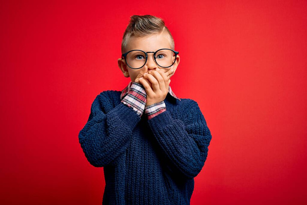 Ein kleines kaukasisches Kind mit blauen Augen, das mit einer intelligenten Brille vor rotem Hintergrund steht und den Mund mit den Händen versehentlich verdeckt. Geheimes Konzept. - Foto, Bild
