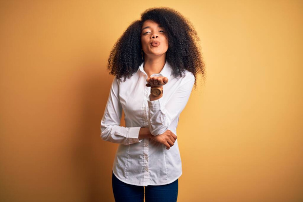 Mladý krásný africký americký elegantní žena s afro vlasy stojící nad žlutým pozadím při pohledu na kameru foukání polibek s rukou na vzduchu je krásné a sexy. Výraz lásky. - Fotografie, Obrázek