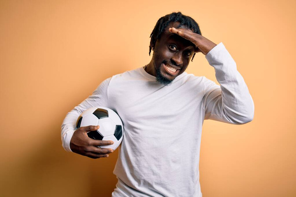 頭に手で強調黄色の背景にサッカーボールを保持サッカーをプレイ若いアフリカ系アメリカ人選手の男は、恥と驚きの顔でショックを受け、怒りと不満。恐怖と過ちへの動揺. - 写真・画像