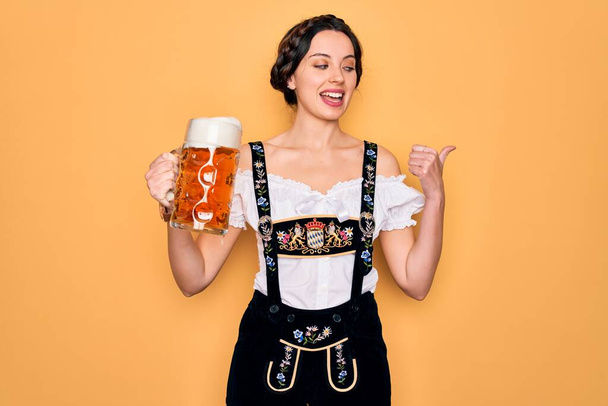 Гарна жінка з блакитними очима, одягнена в традиційне вбрання на октоберфест, яка п'є баночку з написом пива і показує пальцем до сторони з радісним обличчям посміхаючись - Фото, зображення
