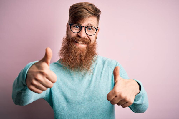 Beau rouquin irlandais avec barbe portant des lunettes sur fond rose isolé signe de succès faire geste positif avec la main, pouces levés souriant et heureux. Expression joyeuse et geste gagnant
. - Photo, image