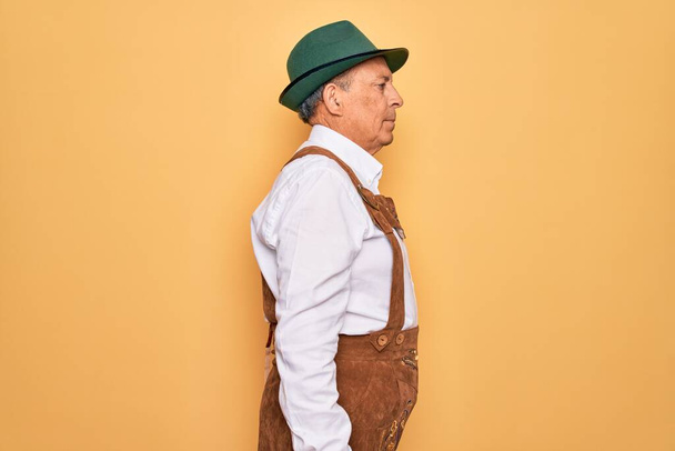 Homme âgé aux cheveux gris portant un costume traditionnel allemand octoberfest sur fond jaune regardant vers le côté, pose de profil relaxante avec un visage naturel et un sourire confiant
. - Photo, image