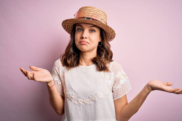Young beautiful blonde girl wearing summer hat over pink isolated background clueless and confused expression with arms and hands raised. Doubt concept. - 写真・画像