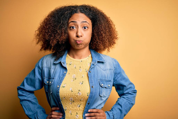 Young african american woman with afro hair wearing casual denim shirt over yellow background puffing cheeks with funny face. Mouth inflated with air, crazy expression. - Fotografie, Obrázek