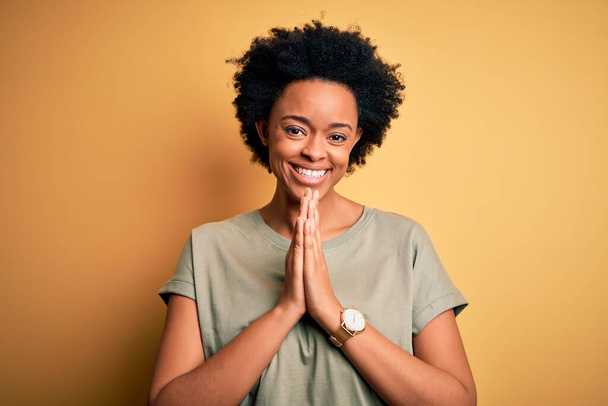 Νεαρή όμορφη Αφρο-Αμερικανίδα με σγουρά μαλλιά φορώντας casual t-shirt προσεύχεται με τα χέρια μαζί ζητώντας συγχώρεση χαμογελώντας σίγουροι. - Φωτογραφία, εικόνα