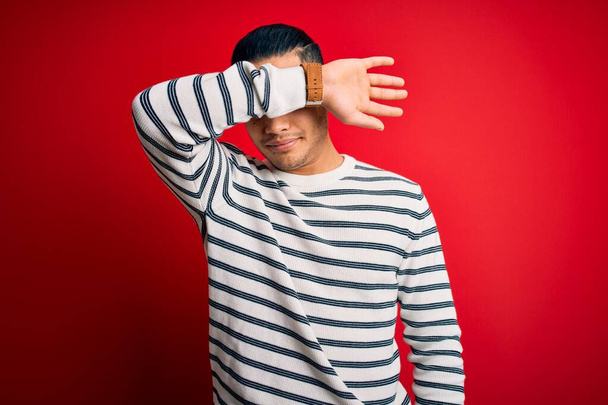 Young brazilian man wearing casual striped t-shirt standing over isolated red background covering eyes with arm, looking serious and sad. Sightless, hiding and rejection concept - Foto, afbeelding