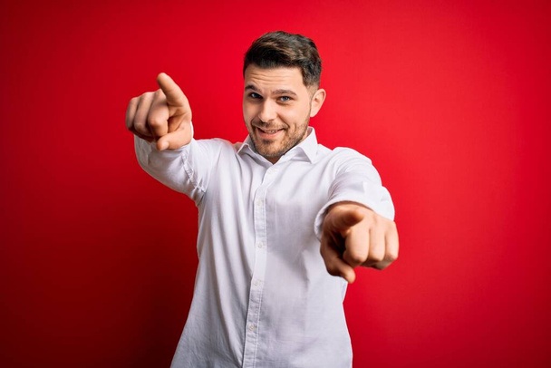 Young business man with blue eyes wearing elegant shirt standing over red isolated background pointing to you and the camera with fingers, smiling positive and cheerful - Photo, Image