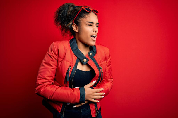 Beautiful african american girl wearing red jacket and sunglasses over isolated background with hand on stomach because nausea, painful disease feeling unwell. Ache concept. - Fotó, kép