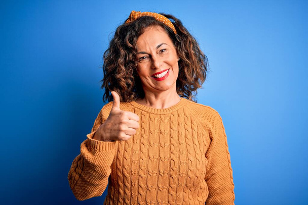 Middle age beautiful woman wearing casual yellow sweater over isolated yellow background doing happy thumbs up gesture with hand. Approving expression looking at the camera showing success. - Photo, Image