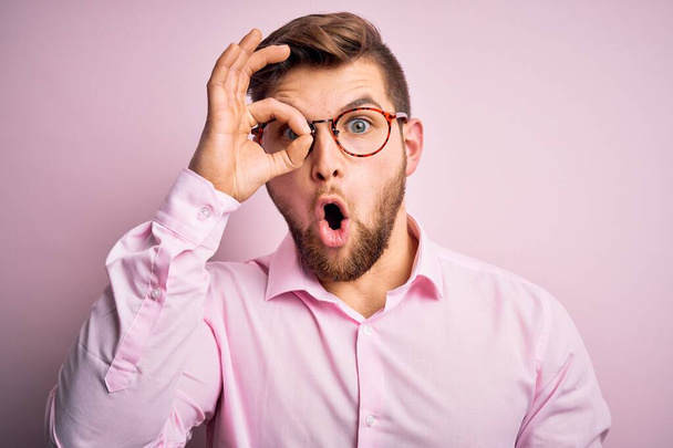 Young handsome blond man with beard and blue eyes wearing pink shirt and glasses doing ok gesture shocked with surprised face, eye looking through fingers. Unbelieving expression. - Photo, Image
