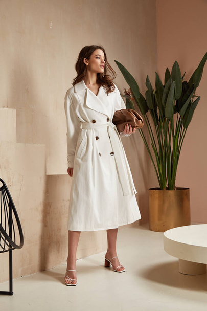 Beautiful brunette woman natural make up wear fashion clothes casual dress code office style white long cloak for romantic date business meeting accessory bag lather interior boho stairs flowerpot. - 写真・画像