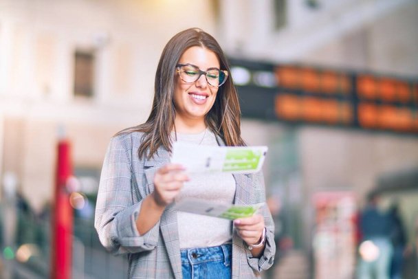Молодая красивая женщина улыбается счастливой и уверенной. Стоя с улыбкой на лице держа билеты на вокзале
 - Фото, изображение