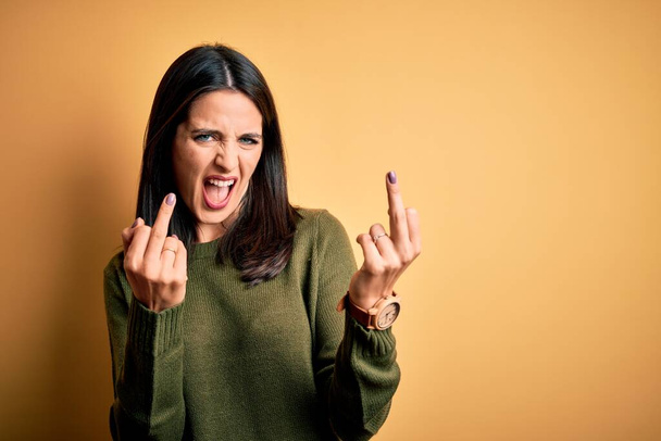 Young brunette woman with blue eyes wearing green casual sweater over yellow background Showing middle finger doing fuck you bad expression, provocation and rude attitude. Screaming excited - Foto, Bild