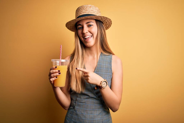Jovem mulher turística bonita de férias usando chapéu de verão bebendo suco de laranja saudável muito feliz apontando com a mão e o dedo
 - Foto, Imagem