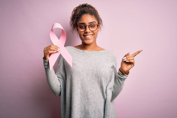 若いですアフリカ系アメリカ人の女の子保持ピンク癌リボン上の隔離された背景非常に幸せなポインティングとともに手と指へ側面 - 写真・画像