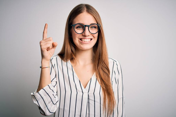 Young beautiful redhead woman wearing casual striped t-shirt and glasses pointing finger up with successful idea. Exited and happy. Number one. - Foto, immagini