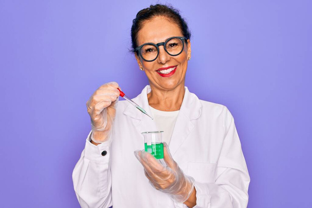 Старша вчена середнього віку жінка в лабораторному пальто тримає дослідницьку пробірку з щасливим обличчям, що стоїть і посміхається з впевненою посмішкою, показуючи зуби
 - Фото, зображення