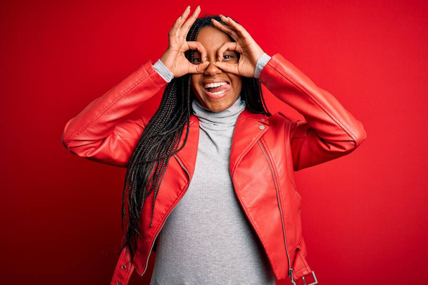 Mujer afroamericana joven que usa una chaqueta de cuero de moda fresca sobre fondo rojo aislado haciendo un gesto aceptable como binoculares sacando la lengua, ojos mirando a través de los dedos. Expresión loca
. - Foto, imagen