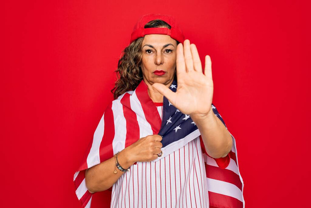 Patriotische Frau mittleren Alters in Baseballausrüstung, die mit offener Hand die USA-Flagge hält und mit ernstem und selbstbewusstem Gesichtsausdruck ein Stoppschild macht, Verteidigungsgeste - Foto, Bild