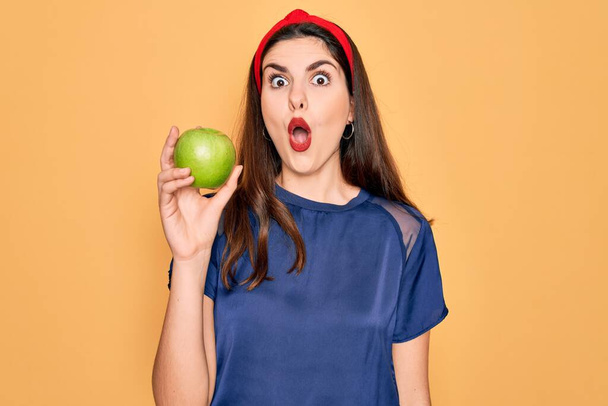 Młoda piękna dziewczyna jedząc świeże organiczne zdrowe zielone jabłko na żółtym tle przestraszony w szoku z twarzy niespodzianka, boi się i podekscytowany ekspresją strachu - Zdjęcie, obraz