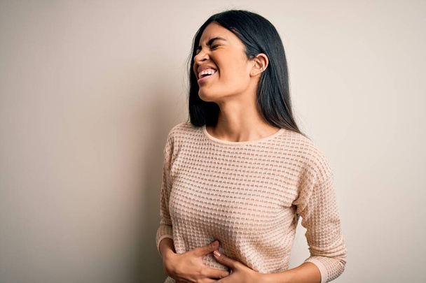 胃の上に手で隔離された背景にエレガントなピンクのセーターを身に着けている若い美しいヒスパニック系の女性は吐き気、痛みを伴う病気が具合が悪い。アチェの概念. - 写真・画像