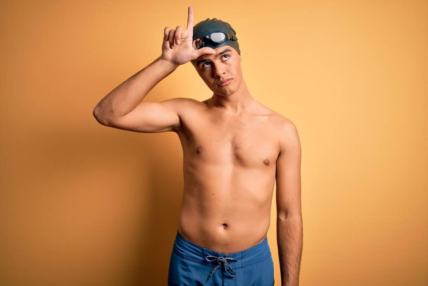 Молодой красивый мужчина без рубашки в купальнике и плавательной шапочке на изолированном желтом фоне высмеивает людей с пальцами на лбу, высмеивая и оскорбляя жесты неудачников.
. - Фото, изображение