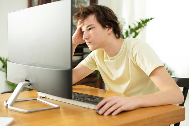 黄色のTシャツを着た若い男がコンピューターの家で働いてる。コロナウイルスの隔離中に隔離に関するインターネット経由の遠隔作業。フリーランサー. - 写真・画像