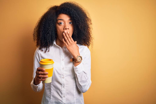 Молодая африканская деловая женщина с афроволосами пьет кофе из чашечки, прикрывая рот рукой, потрясенной стыдом за ошибку, выражением страха, испуганной в тишине, секретной концепцией
 - Фото, изображение