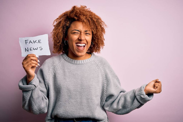 Young African American afro woman with curly hair holding paper with fake news message screaming proud and celebrating victory and success very excited, cheering emotion - Photo, Image
