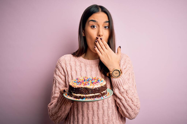 Giovane bella donna bruna che tiene compleanno deliziosa torta su sfondo rosa coprire la bocca con la mano scioccata dalla vergogna per errore, espressione di paura, spaventata in silenzio, concetto segreto
 - Foto, immagini