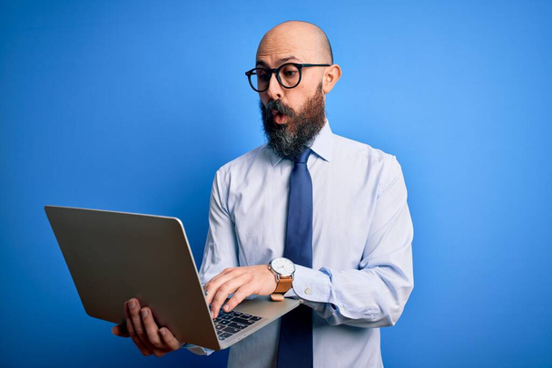 Sakallı yakışıklı bir iş adamı mavi arka planda dizüstü bilgisayar kullanıyor. Şaşırmış bir yüzle korkmuş ve korku ifadesinden heyecanlanmış. - Fotoğraf, Görsel