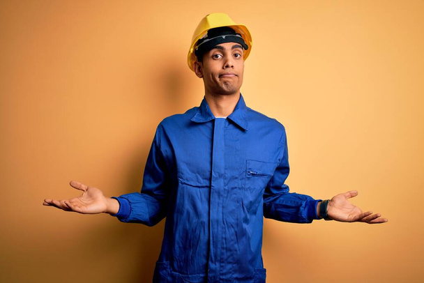 Young handsome african american worker man wearing blue uniform and security helmet clueless and confused with open arms, no idea concept. - Photo, Image