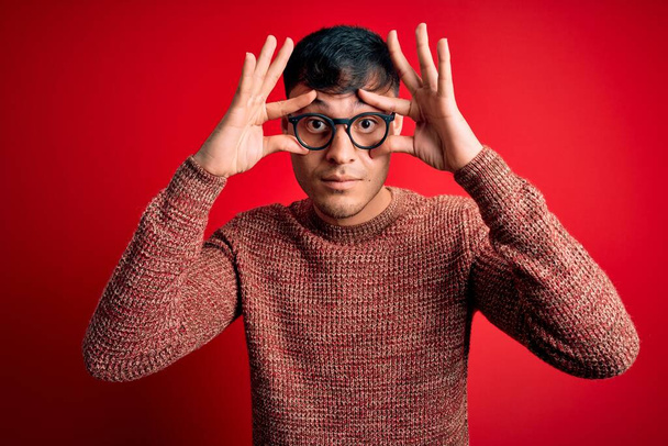 Joven hombre hispano guapo usando anteojos nerd sobre fondo rojo tratando de abrir los ojos con los dedos, soñoliento y cansado para la fatiga de la mañana
 - Foto, Imagen