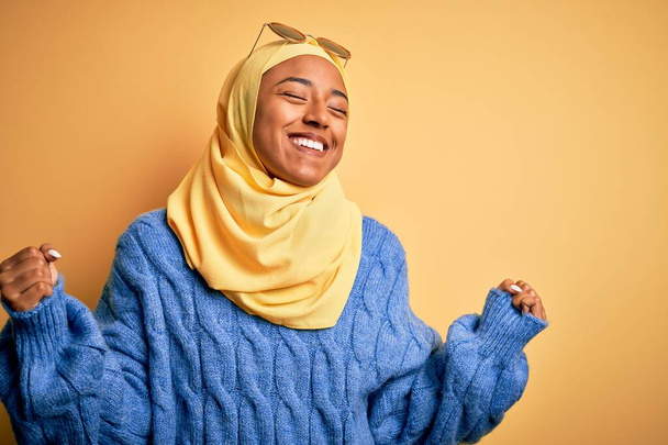 Młoda Afroamerykanka ubrana w żółty hidżab muzułmański i okulary przeciwsłoneczne bardzo szczęśliwa i podekscytowana wykonując gest zwycięzcy z podniesionymi ramionami, uśmiechnięta i krzycząca o sukces. Koncepcja uroczystości. - Zdjęcie, obraz
