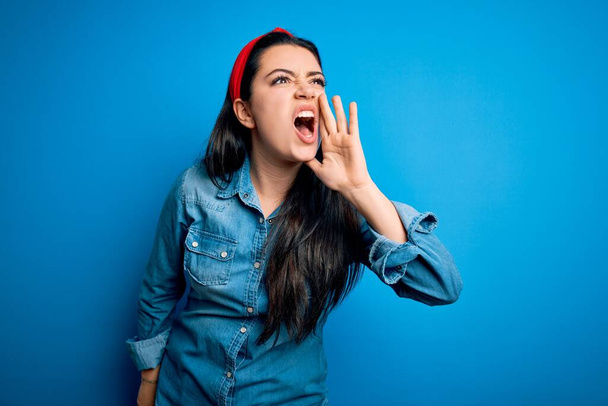Junge brünette Frau in lässigem Jeanshemd über blauem isoliertem Hintergrund schreit und schreit laut Seite an Seite mit der Hand auf dem Mund. Kommunikationskonzept. - Foto, Bild