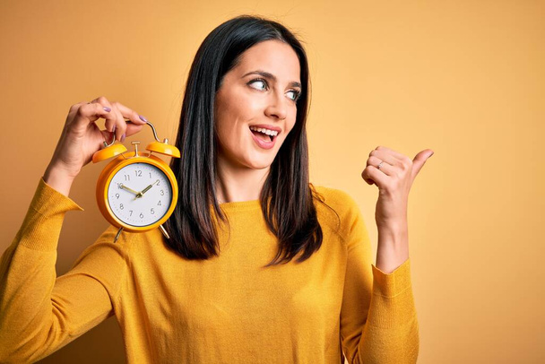 Young woman with blue eyes holding alarm clock standing over isolated yellow background pointing and showing with thumb up to the side with happy face smiling - Photo, Image