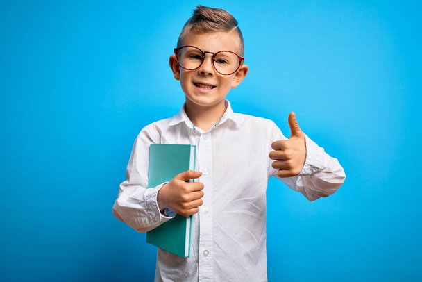 Молодой маленький кавказский студент в умных очках и держит книгу из школы счастливой с большой улыбкой делает хорошо знак, палец вверх пальцами, отличный знак
 - Фото, изображение