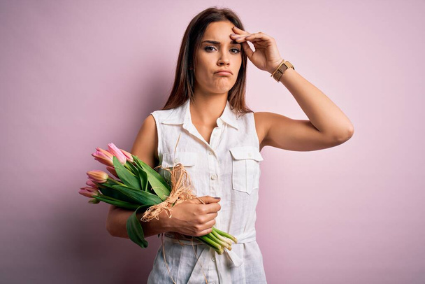 Młoda piękna brunetka trzyma bukiet kwiatów tulipanów na różowym tle zaniepokojony i zestresowany o problem z ręką na czole, nerwowy i zaniepokojony kryzysem - Zdjęcie, obraz