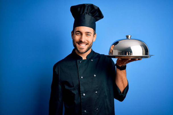 Νεαρός μάγειρας με γενειάδα φοράει ενιαίο δίσκο κρατώντας με τρούλο πάνω από το μπλε φόντο με χαρούμενο πρόσωπο στέκεται και χαμογελά με αυτοπεποίθηση χαμόγελο που δείχνει τα δόντια - Φωτογραφία, εικόνα