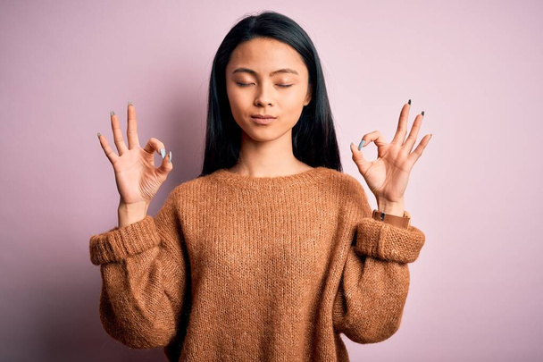 Young beautiful chinese woman wearing casual sweater over isolated pink background relax and smiling with eyes closed doing meditation gesture with fingers. Yoga concept. - Fotoğraf, Görsel