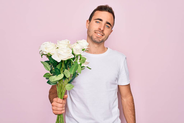 Joven hombre guapo con ojos azules sosteniendo ramo de flores sobre fondo rosa con una cara feliz de pie y sonriendo con una sonrisa confiada mostrando los dientes
 - Foto, imagen