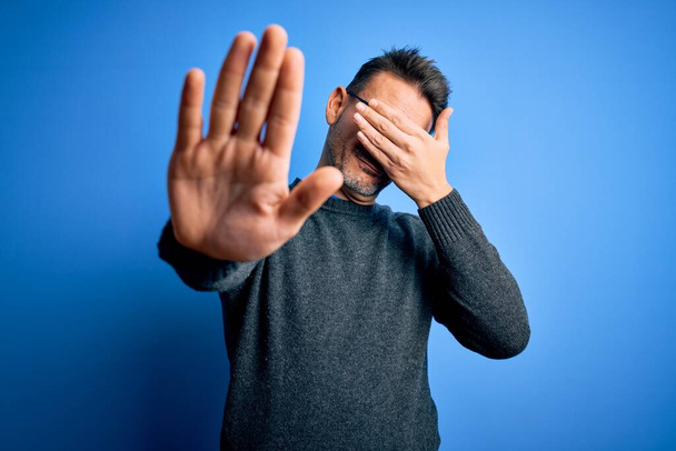 Young handsome man wearing casual sweater and glasses standing over blue background covering eyes with hands and doing stop gesture with sad and fear expression. Embarrassed and negative concept. - Photo, Image