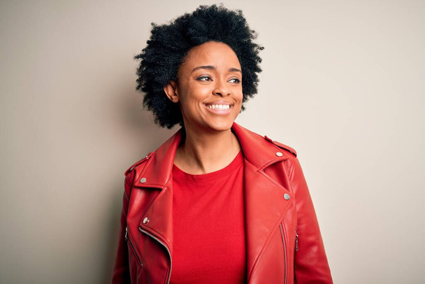 Jonge mooie Afro-Amerikaanse afro vrouw met krullend haar dragen casual rode jas weg te kijken naar kant met glimlach op het gezicht, natuurlijke expressie. Lachend vol vertrouwen. - Foto, afbeelding