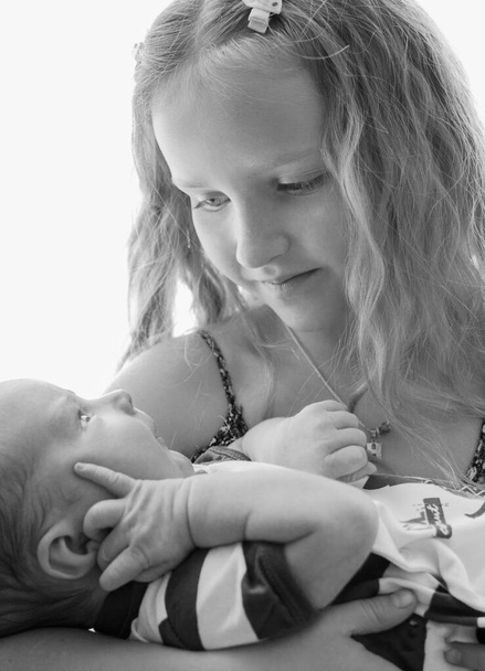 Loving older sister blonde sweetheart touching and gentle holds her newborn brother in her arms, cute tiny baby. The concept of a happy family. Beautiful conceptual of love, trust and tenderness - Fotoğraf, Görsel