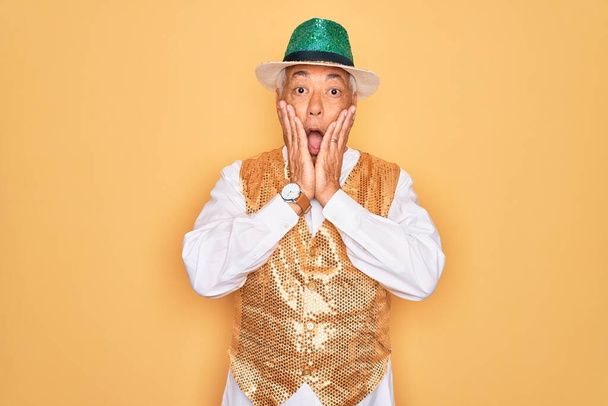 Homem de cabelos grisalhos sênior de meia idade vestindo carnaval brasileiro custome sobre fundo amarelo com medo e chocado, surpresa e expressão espantada com as mãos no rosto
 - Foto, Imagem