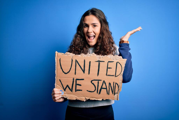 Krásná aktivistka drží prapor s jednotným stojanem poselství nad modrým pozadím velmi šťastný a vzrušený, vítězný výraz slaví vítězství křičí s velkým úsměvem a zvedl ruce - Fotografie, Obrázek