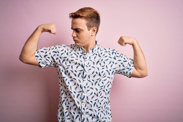 Joven hombre pelirrojo guapo con camisa de verano casual de pie sobre fondo rosa mostrando los músculos de los brazos sonriendo orgulloso. Concepto Fitness
. - Foto, Imagen